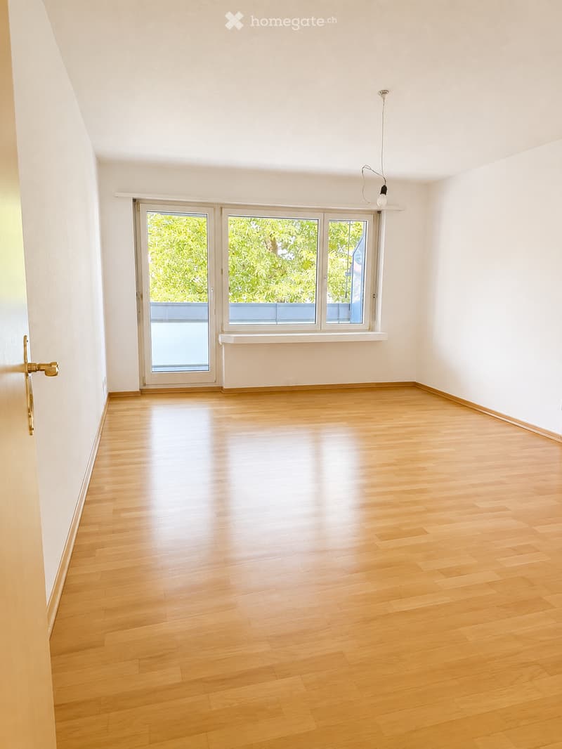 Empty room with wooden flooring, white walls, large windows, and a sliding door leading to a balcony.