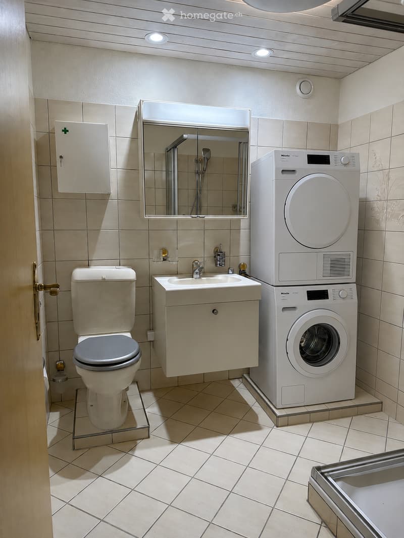 Modern bathroom with beige tiles, a toilet, a sink with a cabinet, a mirror, and a stacked washer/dryer unit.