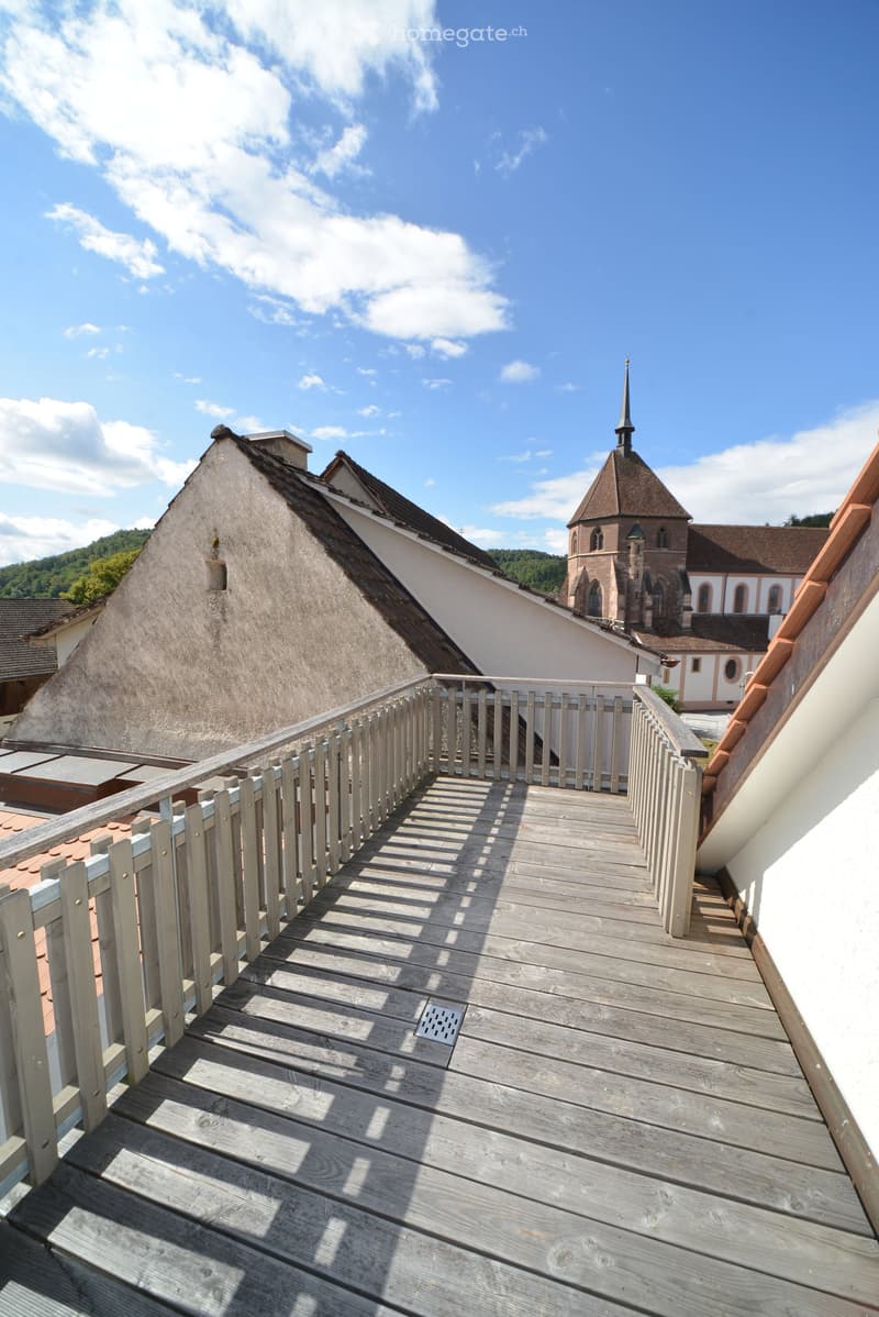 Wooden balcony with white railing, overlooking a historic building with a spire and surrounding rooftops.