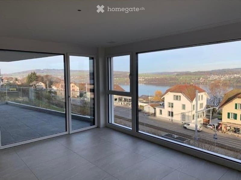 Bright, empty room with large floor-to-ceiling windows offering a scenic view of a lake and surrounding landscape. The space features a modern design with a tiled floor and a glass balcony door.