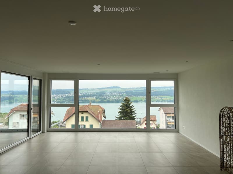 Empty room with large windows offering a scenic view of a lake and surrounding landscape. The room features tiled flooring and modern recessed lighting.