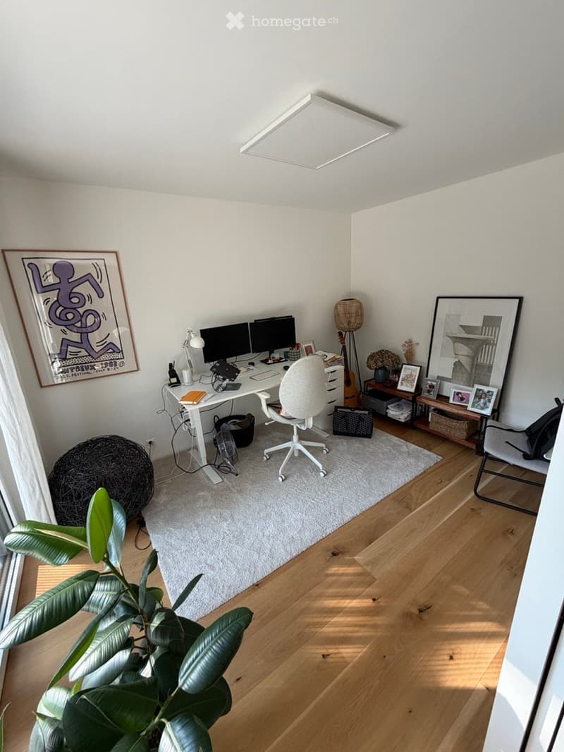 A modern, well-lit home office with a white desk, ergonomic chair, dual monitors, and a variety of decorative items including a guitar, a large plant, and framed artwork.