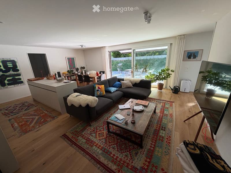 Modern living room with a dark grey sectional sofa, wooden coffee table, laptop, books, and a kitchen area visible in the background. The room features wooden flooring, colorful rugs, and large windows with a view of a garden.