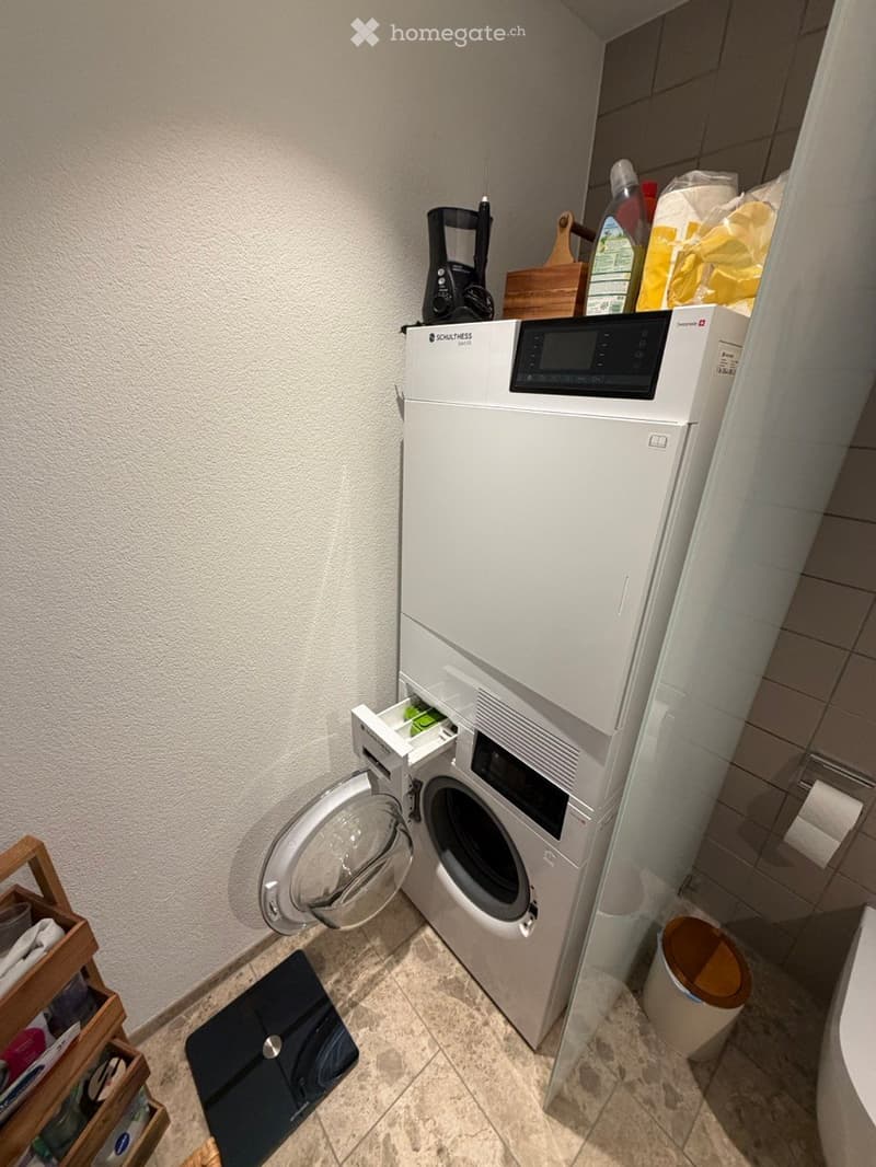 A compact bathroom with a white washing machine and dishwasher unit, a wooden shelf holding cleaning supplies, a trash bin, and a toilet paper holder.