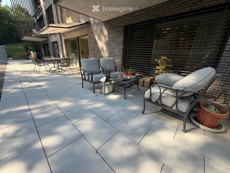 A spacious outdoor patio with tiled flooring, lounge chairs, a coffee table, and a lantern, surrounded by brick walls and greenery.