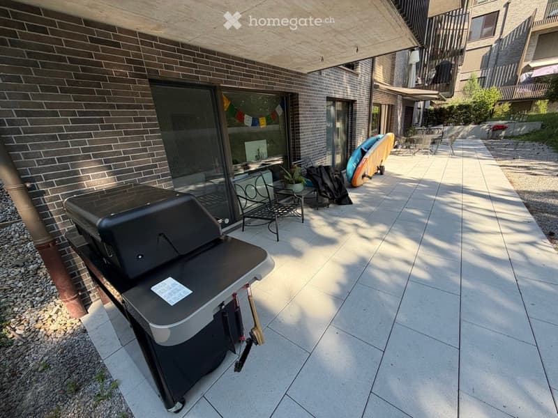 Outdoor patio with a modern grill, chairs, a small table, and surfboards leaning against the wall.
