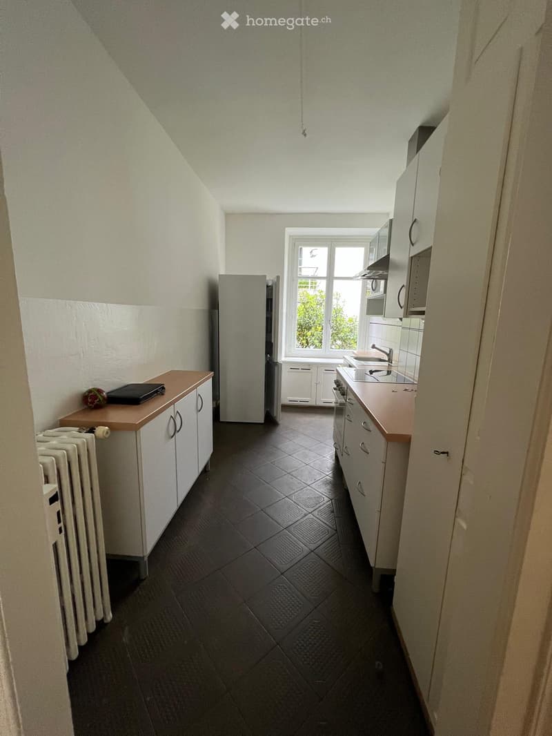 A small, well-lit kitchen with white cabinets, a refrigerator, a sink, and a radiator. The space features a tiled floor and a window letting in natural light.