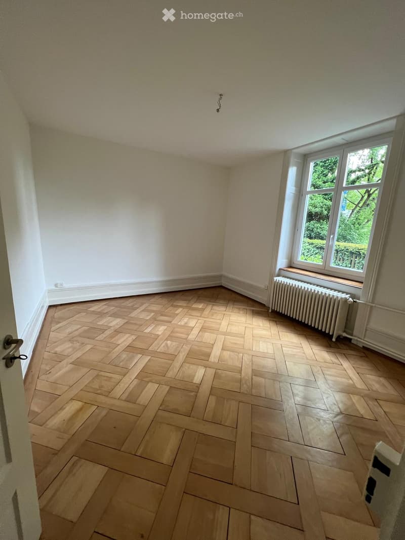 Empty room with wooden herringbone flooring, white walls, and a window with a radiator below it.