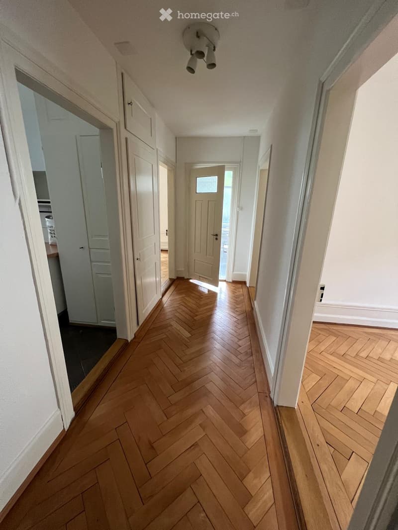 A narrow hallway with herringbone-patterned wooden flooring, white walls, and several doors leading to different rooms.