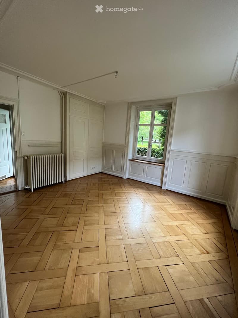 Empty room with wooden herringbone flooring, white walls, a window with a view of greenery, a radiator, and a closed door.