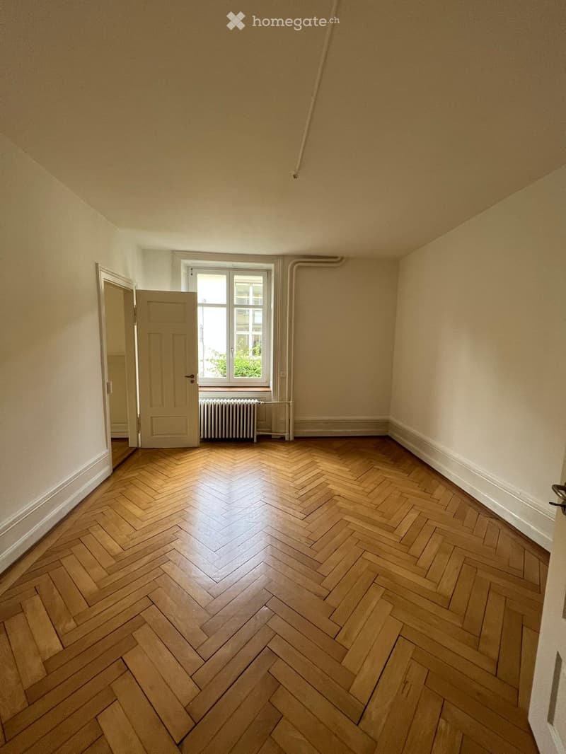 Empty room with wooden herringbone flooring, light-colored walls, a window with white frame, and a radiator below it. A door is visible on the left side.