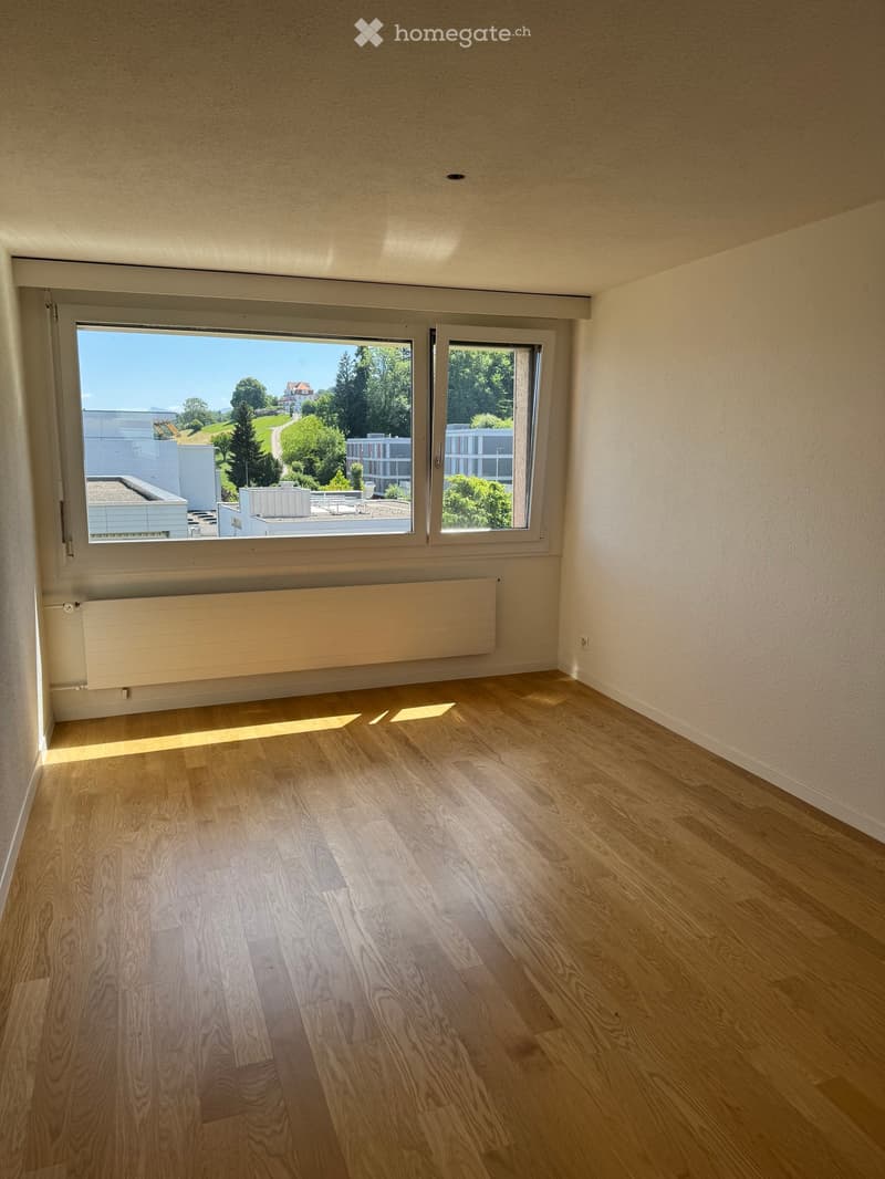 Empty room with wooden flooring, large windows, and a radiator under one window. The view shows greenery and buildings outside.