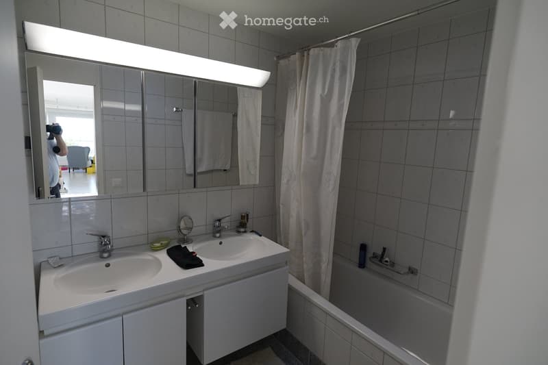 Modern bathroom with double sink, large mirror with built-in lighting, bathtub with shower curtain, and towel on the counter.