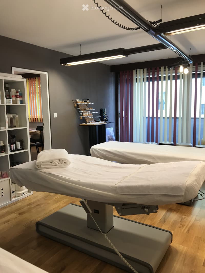 Modern spa treatment room with two treatment tables, shelves with bottles and products, a chair, and a mirror. Large windows with vertical blinds provide natural light.