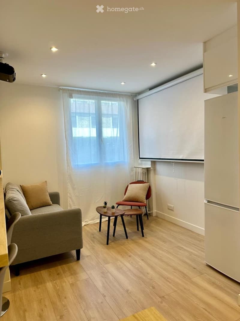 Modern living room with light wooden flooring, a grey sofa with beige cushions, a small coffee table, a red chair, and a large screen mounted on the wall.