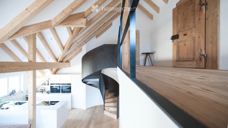 Open-plan hallway with exposed wooden beams, a modern spiral staircase, and a view into a kitchen area with white cabinets and stainless steel appliances.