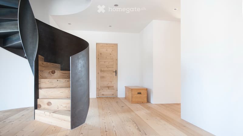 Modern hallway with a wooden floor, a curved metal staircase, a wooden door, and a small wooden chest.