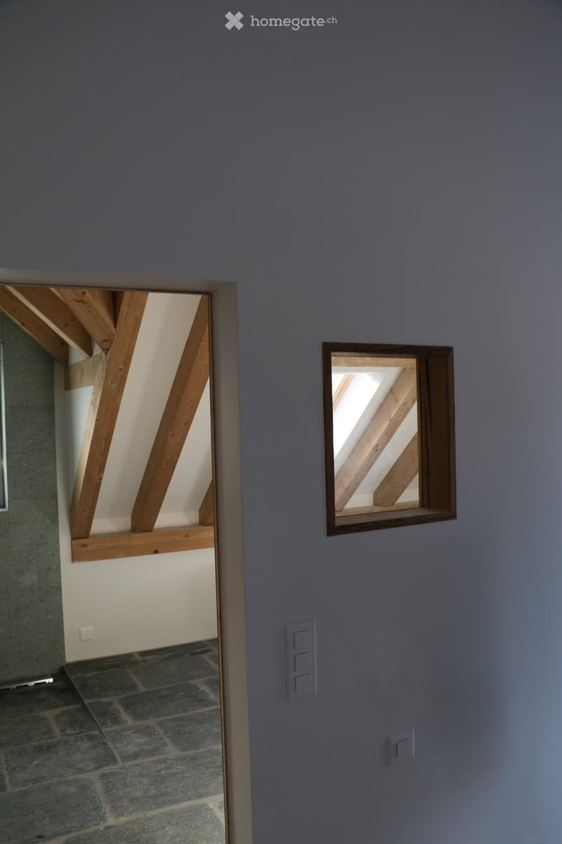 A modern hallway with grey walls, exposed wooden beams visible through a window, and a stone-tiled floor. The hallway features electrical outlets and a small window with wooden framing.