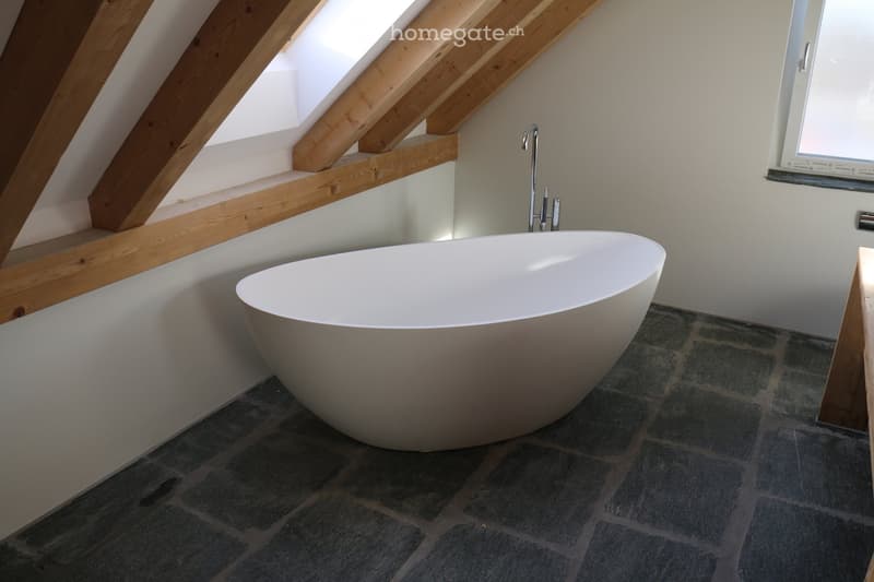 Modern bathroom with a freestanding bathtub, sleek faucet, and exposed wooden beams in the ceiling. The room features a dark tiled floor and a window with blinds.