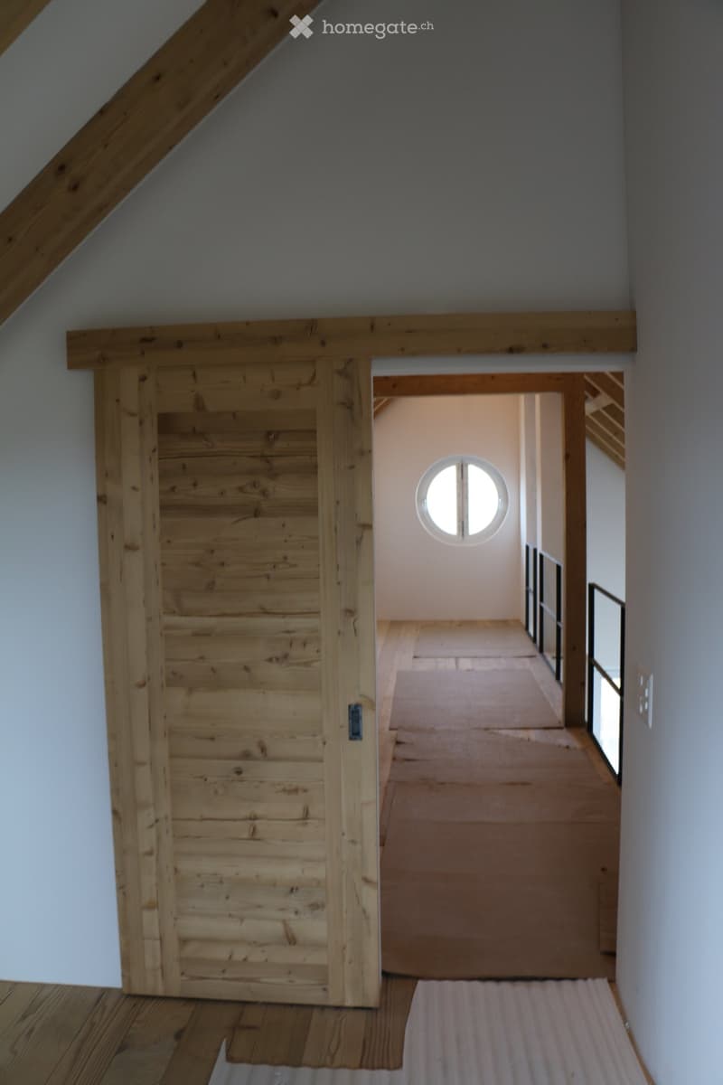 Wooden sliding door leads to a hallway with wooden flooring and a round window at the far end.