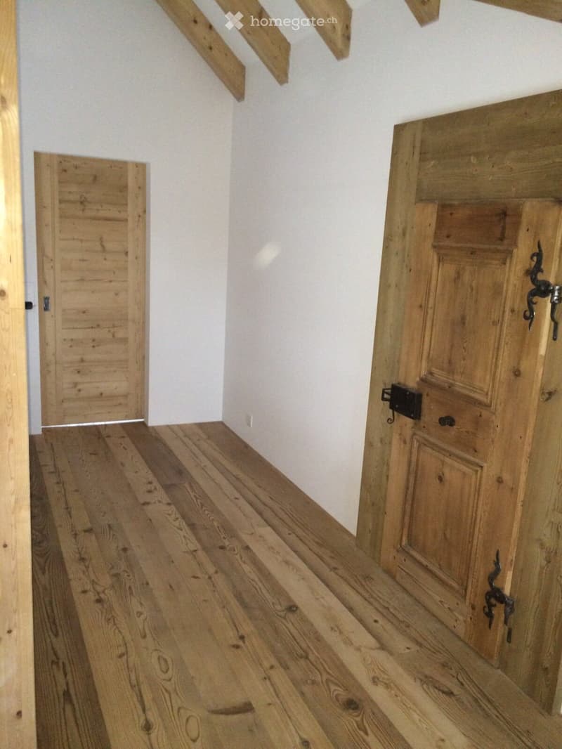 A narrow hallway with wooden floors and white walls, featuring two wooden doors on either side and a staircase leading upwards.