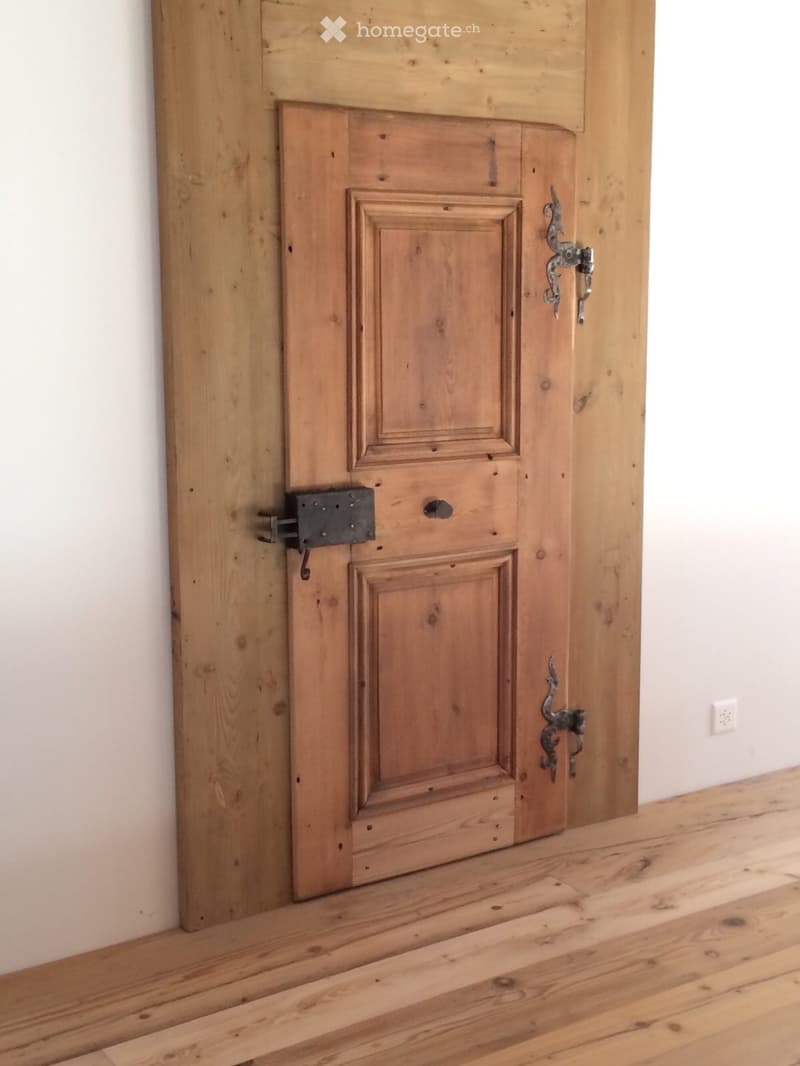 A wooden door with ornate metal handles and a latch, set in a wooden-framed doorway with light-colored walls and a wooden floor.