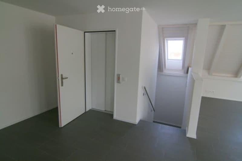 Empty room with white walls, dark tiled floor, open doorway, and a small window. No furniture or equipment visible.
