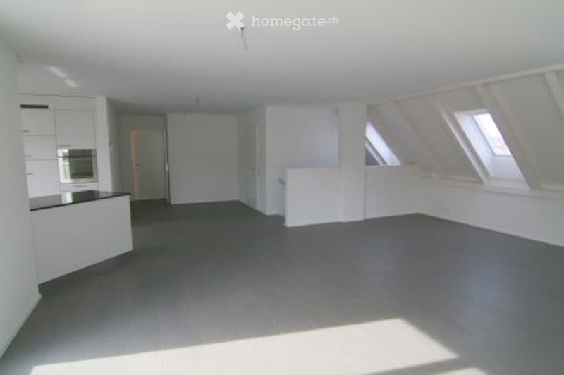Bright, empty attic room with white walls, grey flooring, and skylight windows. Minimalist design with no furniture.