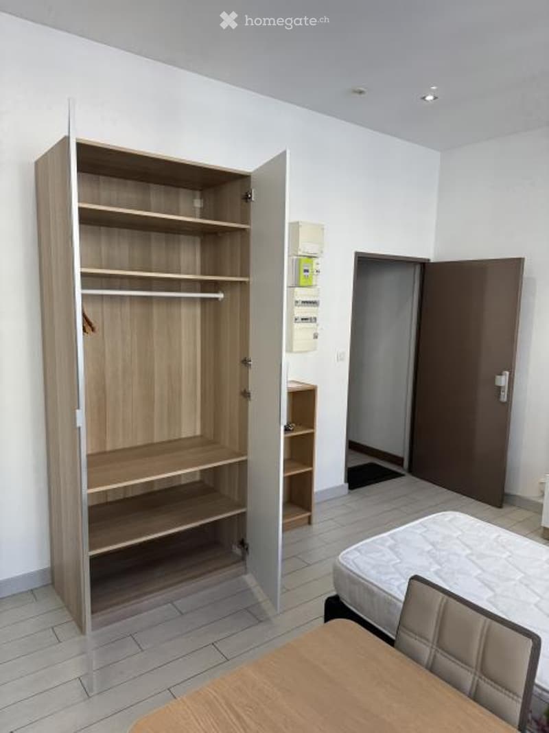 A furnished room with a wooden wardrobe, a bed, a small table, and a chair. The room has white walls and tiled flooring.