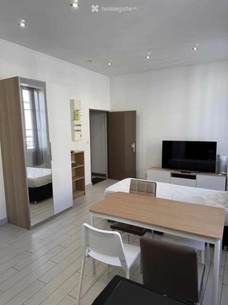 A modern, well-lit room with a large mirror, wardrobe, dining table with chairs, and a television. The space is clean and organized, with a bed visible in the background.