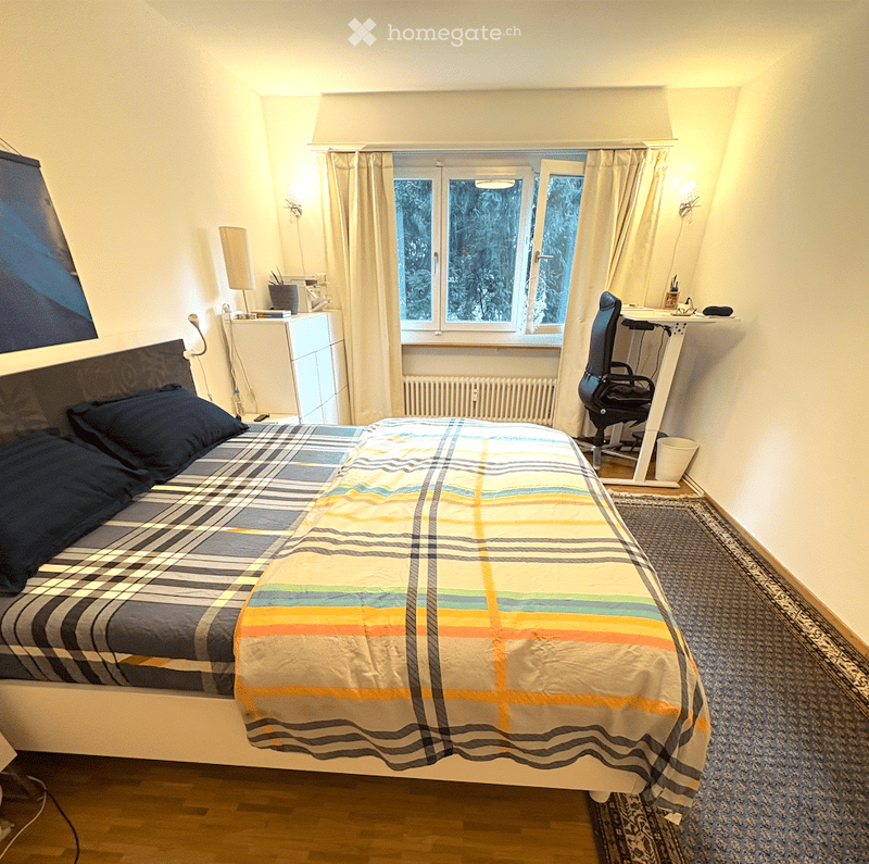Cozy bedroom with a double bed, plaid blanket, desk with chair, window with curtains, and a small nightstand.