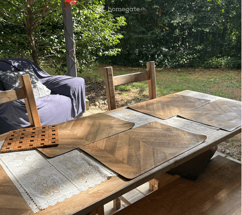 An outdoor patio area with a wooden table, wooden chairs, a woven basket, and a covered sofa in the background.