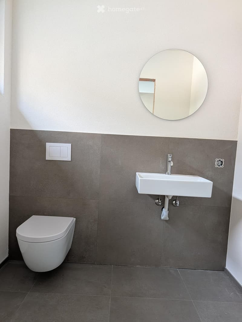 Modern bathroom with a white toilet, a white sink with a chrome faucet, and a round mirror above the sink. The walls are tiled in a two-tone design with light grey upper tiles and dark grey lower tiles.