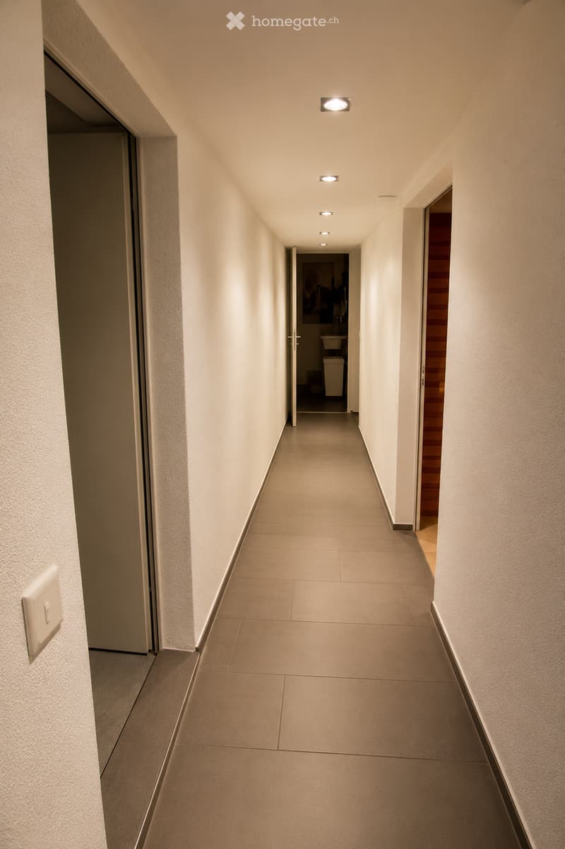 Clean, modern hallway with beige tiled flooring and white walls, featuring recessed ceiling lights and a visible bathroom at the end.