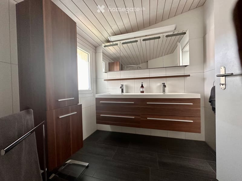 Modern bathroom with a double sink, sleek wooden cabinet, large mirror with integrated lighting, and a small bottle on the counter.