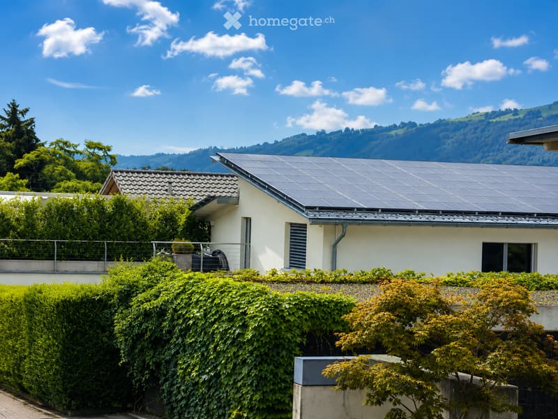 A modern single-story house with solar panels on the roof, surrounded by lush greenery and a well-maintained hedge. The house features a white exterior and a balcony with a metal railing.