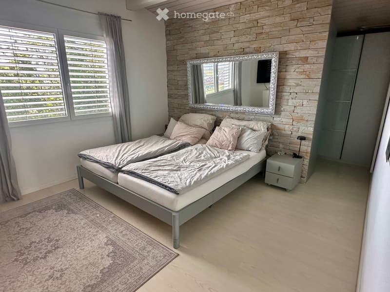 Modern bedroom with a large bed, nightstand, mirror, and a large window with blinds. The walls are decorated with a stone-like texture, and the floor is light-colored wood.