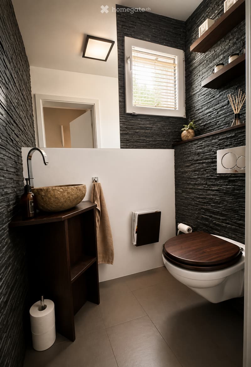 Modern bathroom with dark stone walls, a wooden toilet seat, a stone sink, and a towel hanging on a rack. The room features a window with blinds and a shelf with decorative items.
