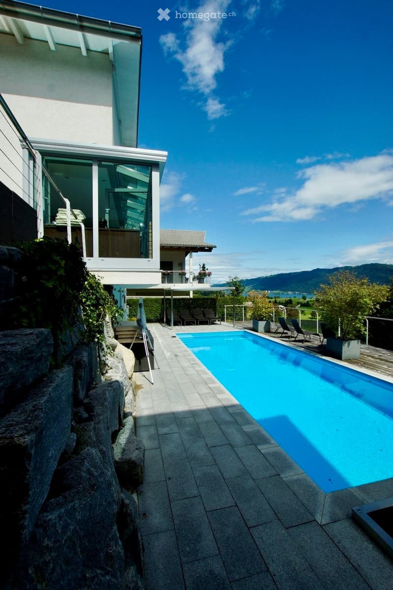 Outdoor area with a swimming pool, lounge chairs, and a modern house in the background, surrounded by greenery and a scenic mountain view.