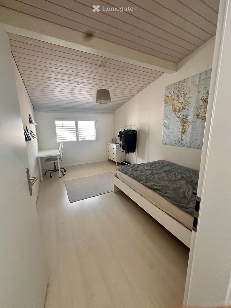Bright bedroom with a single bed, desk, chair, clothing rack, and a world map on the wall. The room features a slanted wooden ceiling and large windows with blinds.