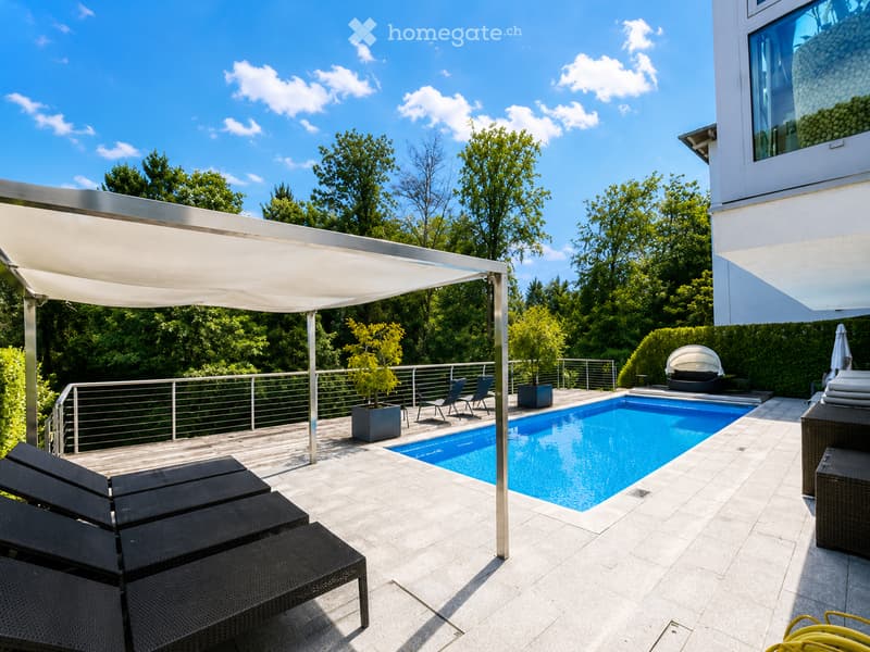 Modern outdoor patio with a swimming pool, lounge chairs, a pergola, and surrounding greenery.