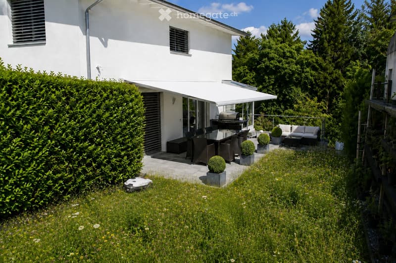 A modern outdoor patio area attached to a white house, featuring a covered seating area with a sofa, coffee table, and chairs, a grill, and several potted plants. The area is surrounded by a neatly trimmed hedge and lush greenery.