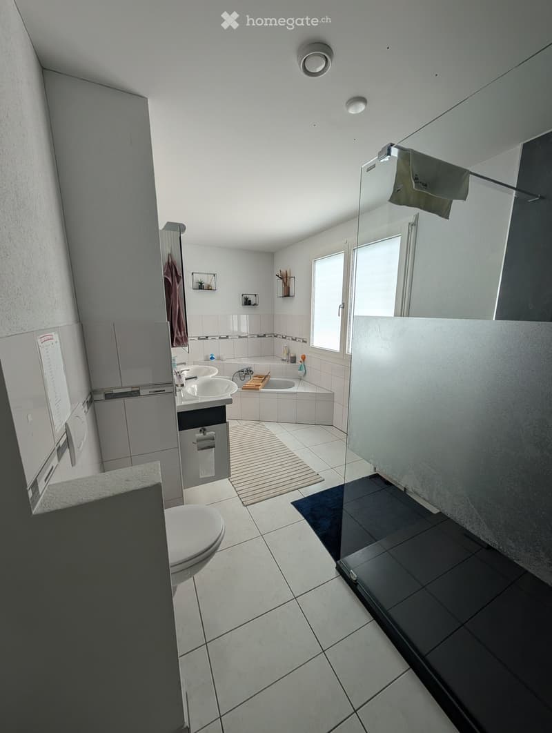 Modern bathroom with white tiles, a bathtub, two sinks, a toilet, and a large mirror. The space includes a shower area with a glass enclosure and a small window for natural light.