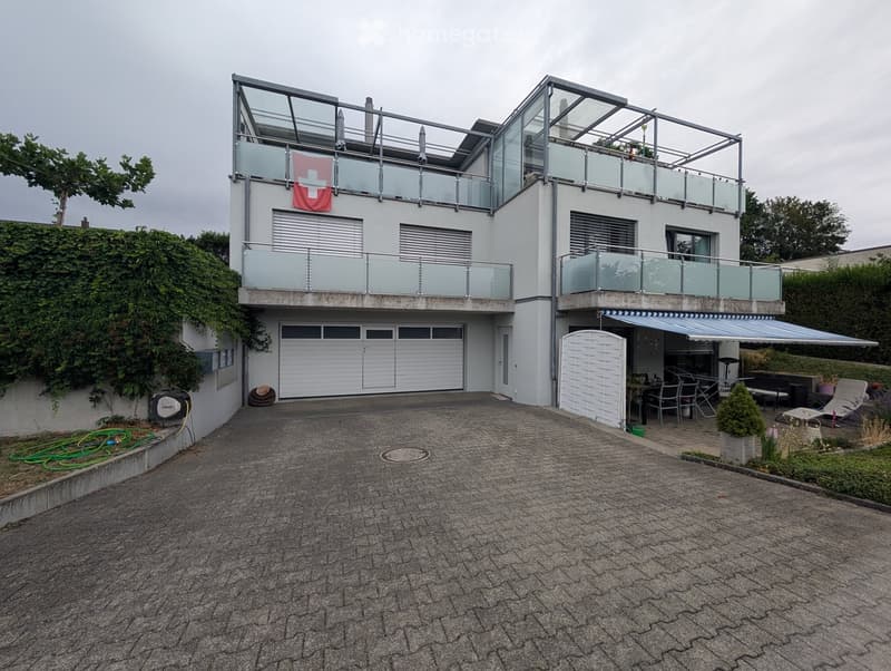 Modern two-story house with white exterior, glass balconies, garage, and outdoor seating area.