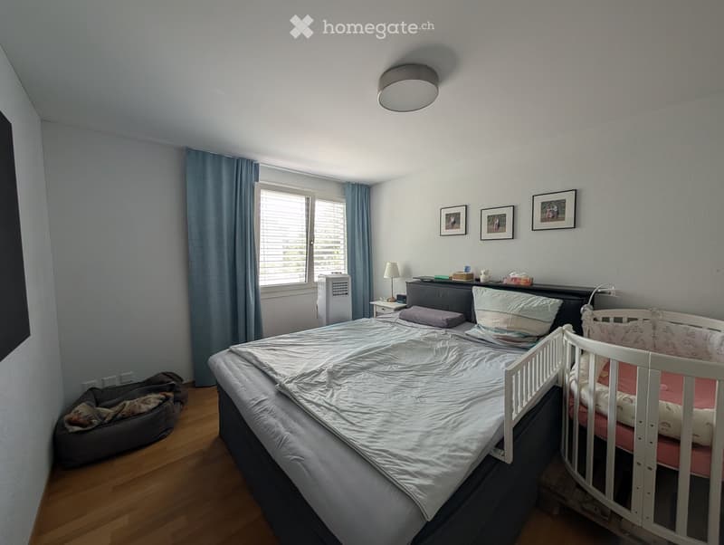 Cozy bedroom with a large bed, a crib, a dog bed, a nightstand with a lamp, an air conditioner, and picture frames on the wall.