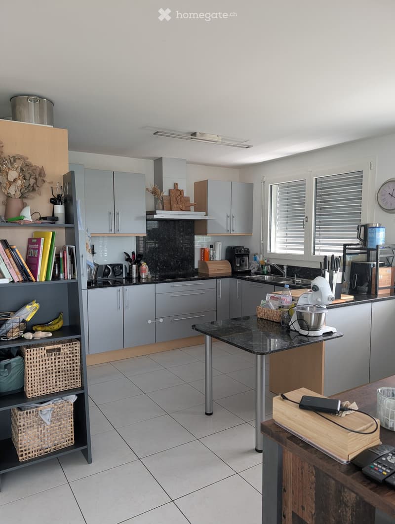 Modern kitchen with grey cabinets, black countertops, and various appliances including a coffee maker, blender, and microwave. There is a bookshelf with books and baskets, and a central island with a black countertop.