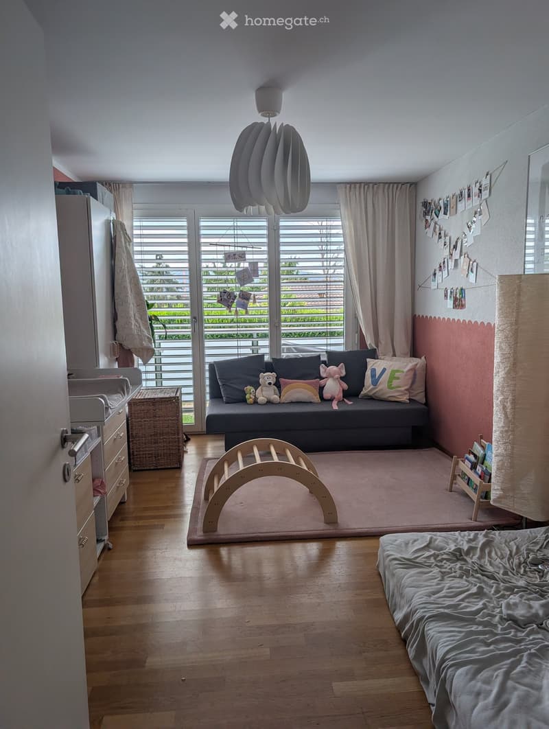 A cozy, well-lit room with a sofa, bed, wooden arch toy, wicker basket, and bookshelf. The room features a large window with blinds and a view of greenery outside.