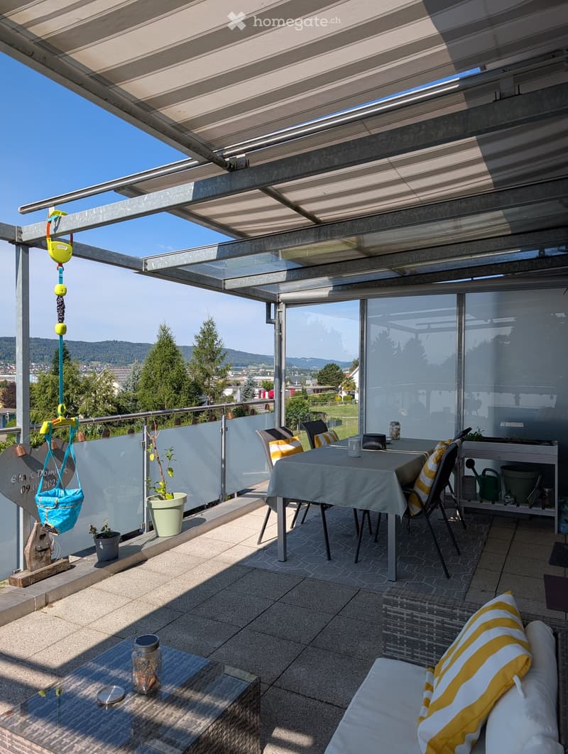 Balcony with a covered area, featuring a dining table with chairs, a sofa with cushions, potted plants, and a hanging swing.