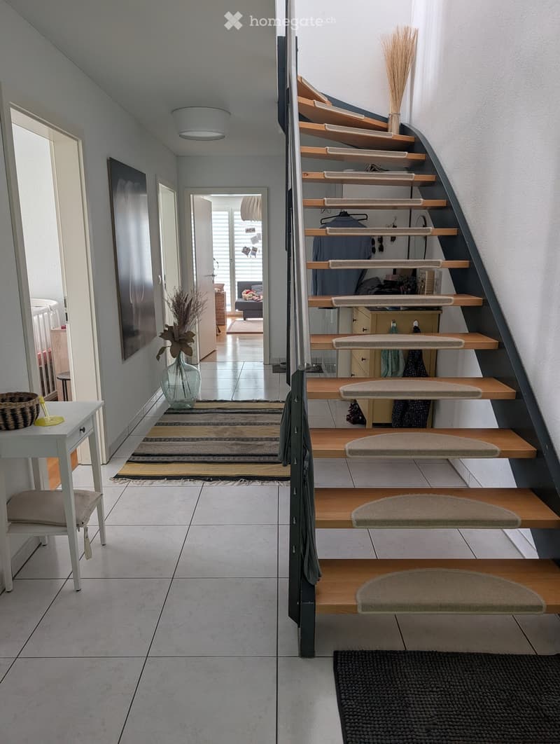 Modern hallway with a curved wooden staircase, tiled floor, and various decorative items including a vase with dried flowers, a small table with a chair, and a woven basket.