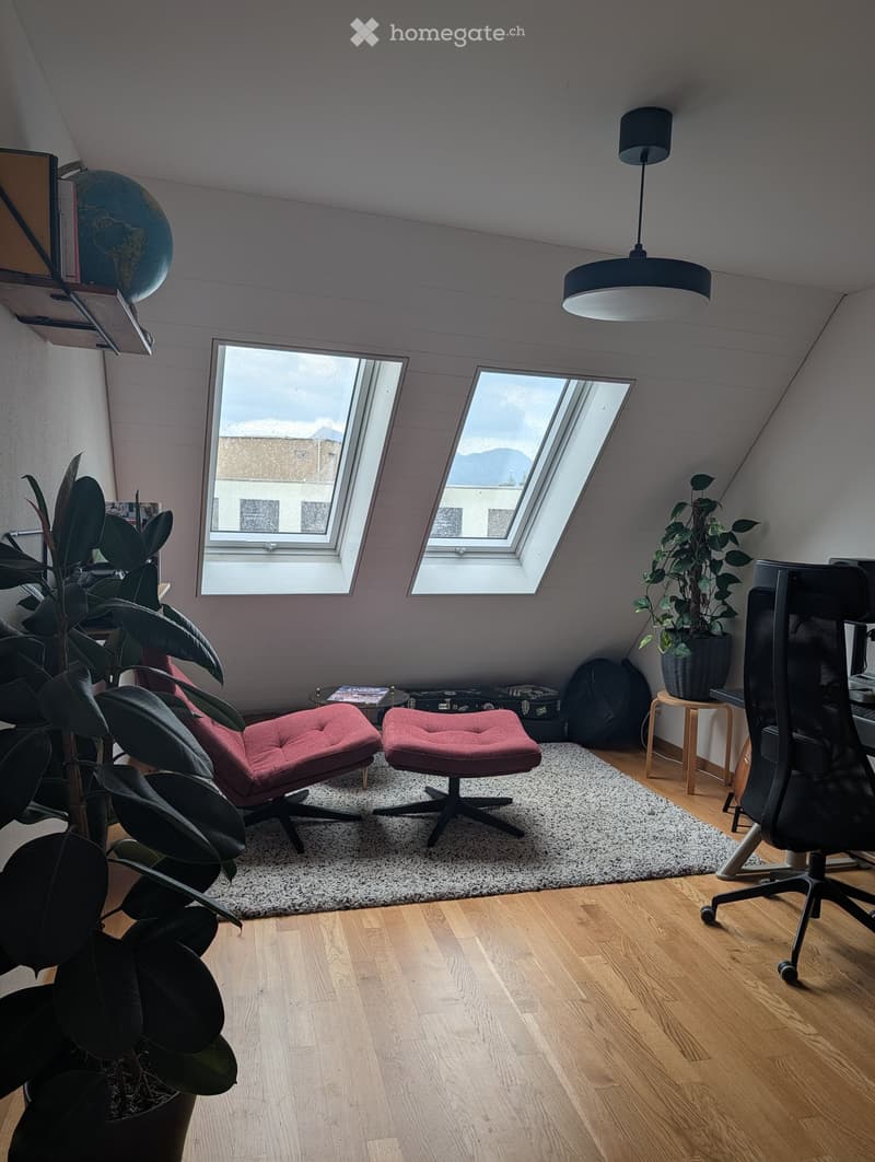 A cozy attic room with wooden floors, skylights, and modern furniture including two red chairs, a small table, a hanging lamp, and a globe on a shelf.